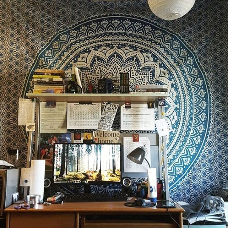 A desk topped with lots of papers, books, supplies, etc. in front of a wall tapestry hung in a dorm room