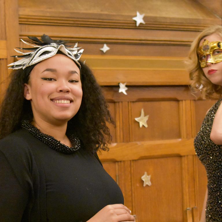 Two female students in formal attire and masquerade masks pose smiling